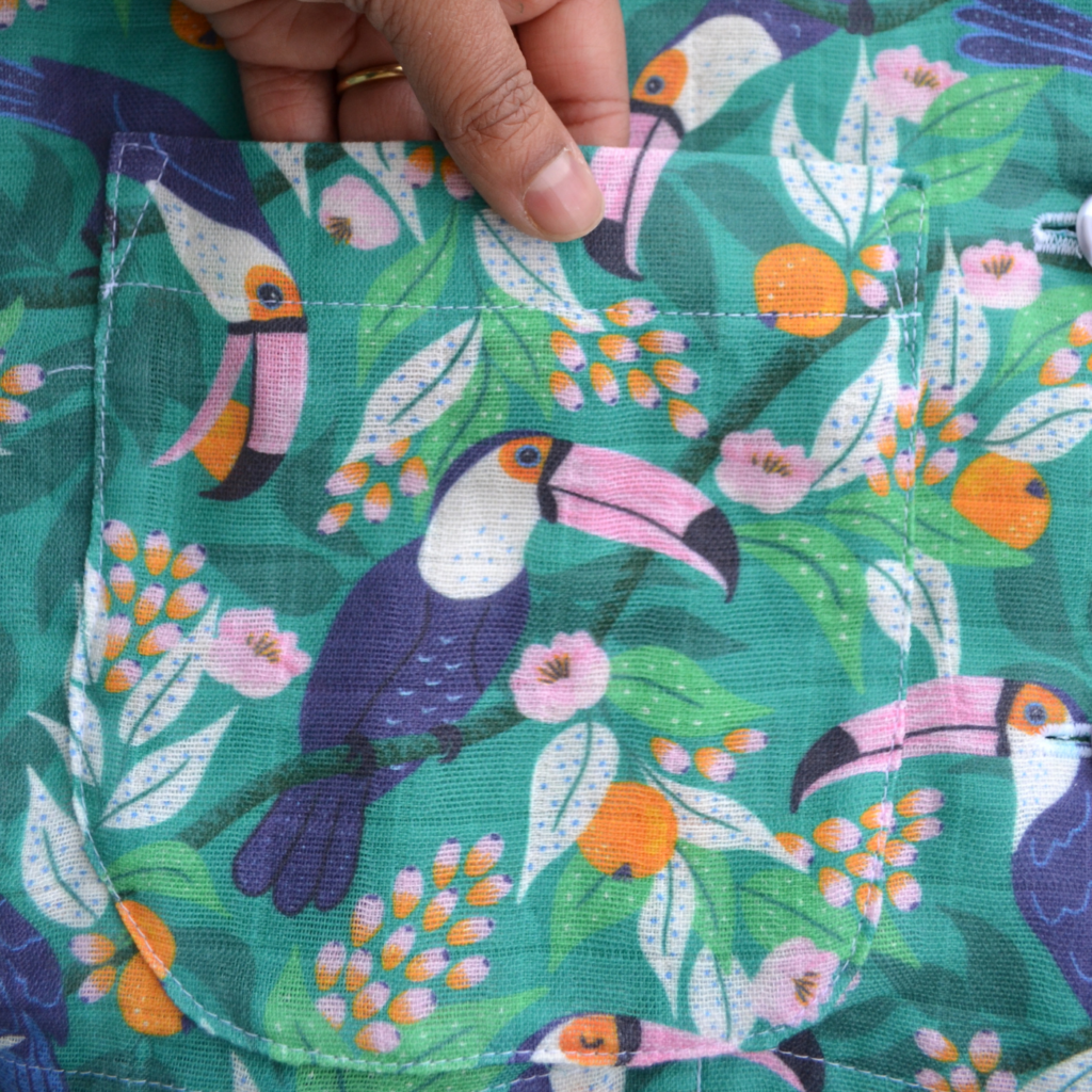 A close up of a front pocket of pajamas with a tropical print featuring toucans, pink and orange flowers and a bright blue background.