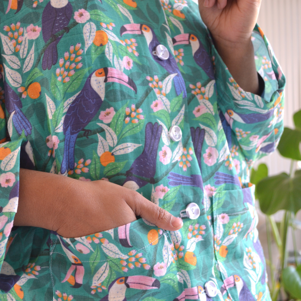 A close up of Rumana’s pajamas showing the top’s front buttons and pockets. Rumana’s left hand is in her pocket. The pajamas have a tropical print featuring toucans, pink and orange flowers and a bright blue background. Behind Rumana is a gray couch and a monstera plant in a raised light bright wooden planter. 