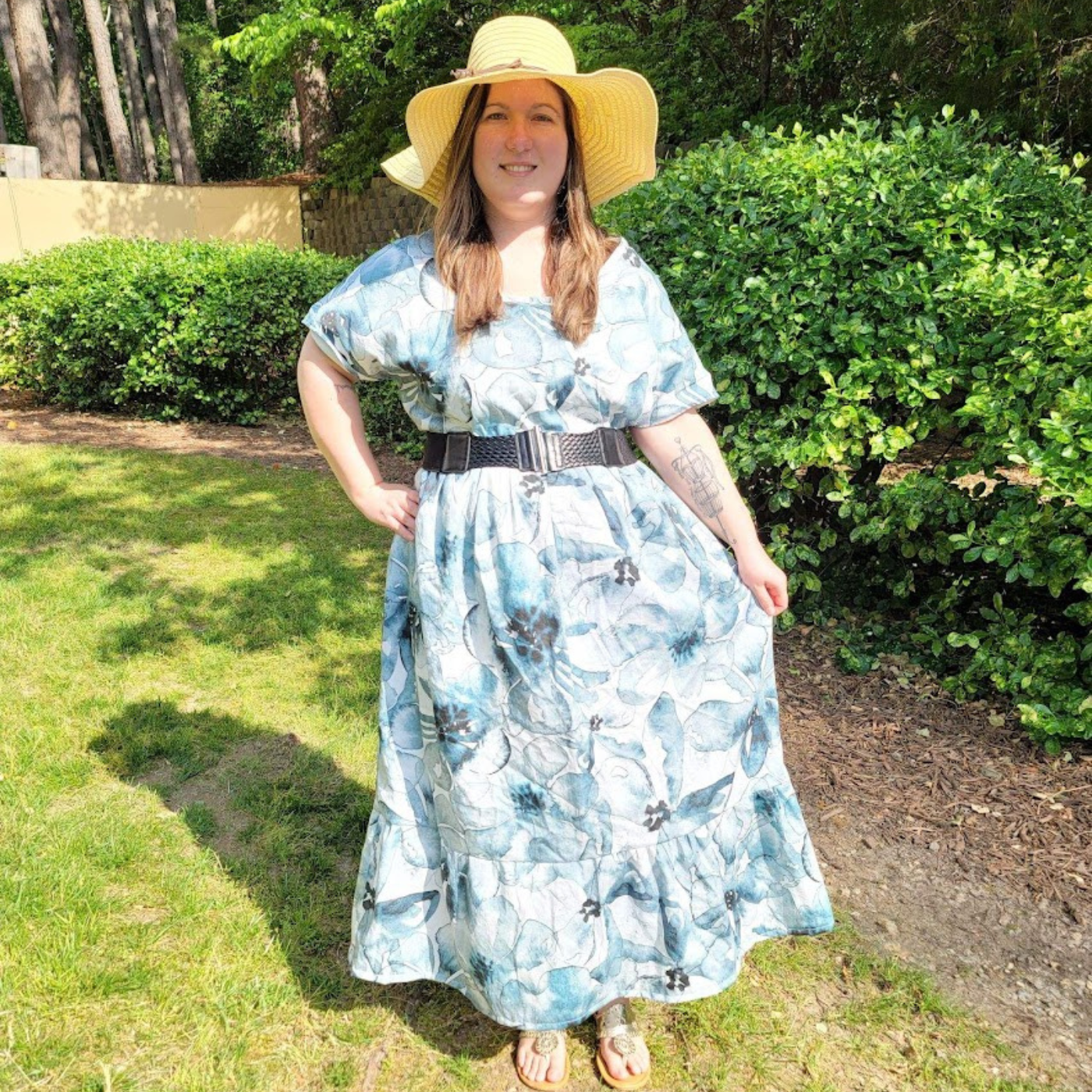 Sarah stands in the grass, smiling at the camera. She is wearing a light brown sunhat and a long dress with a black belt at the waist. The dress has a white background and a watercolor blue flower design. The flowers have black centers.