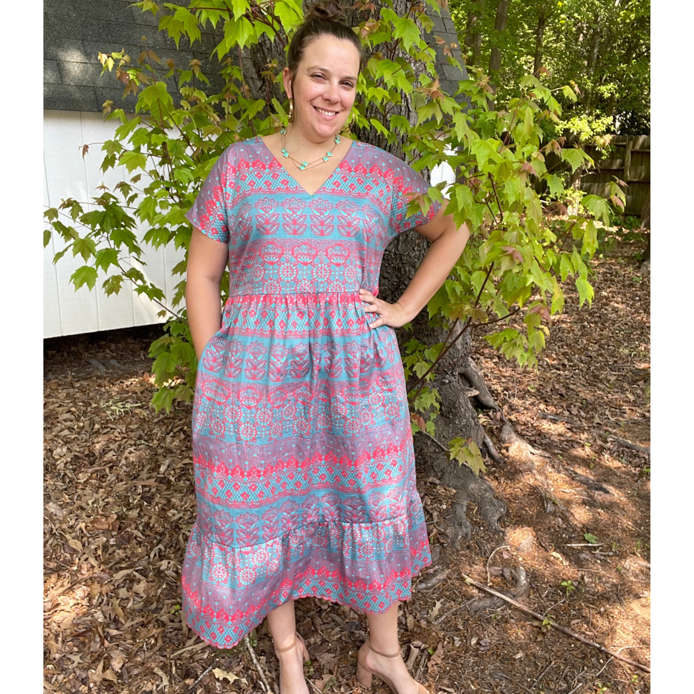Sarah stands with her left hand on her hip and her right hand in her pocket in the mulch in a backyard around some trees. She is smiling at the camera and wearing a long turquoise dress with a hot pink lace design.