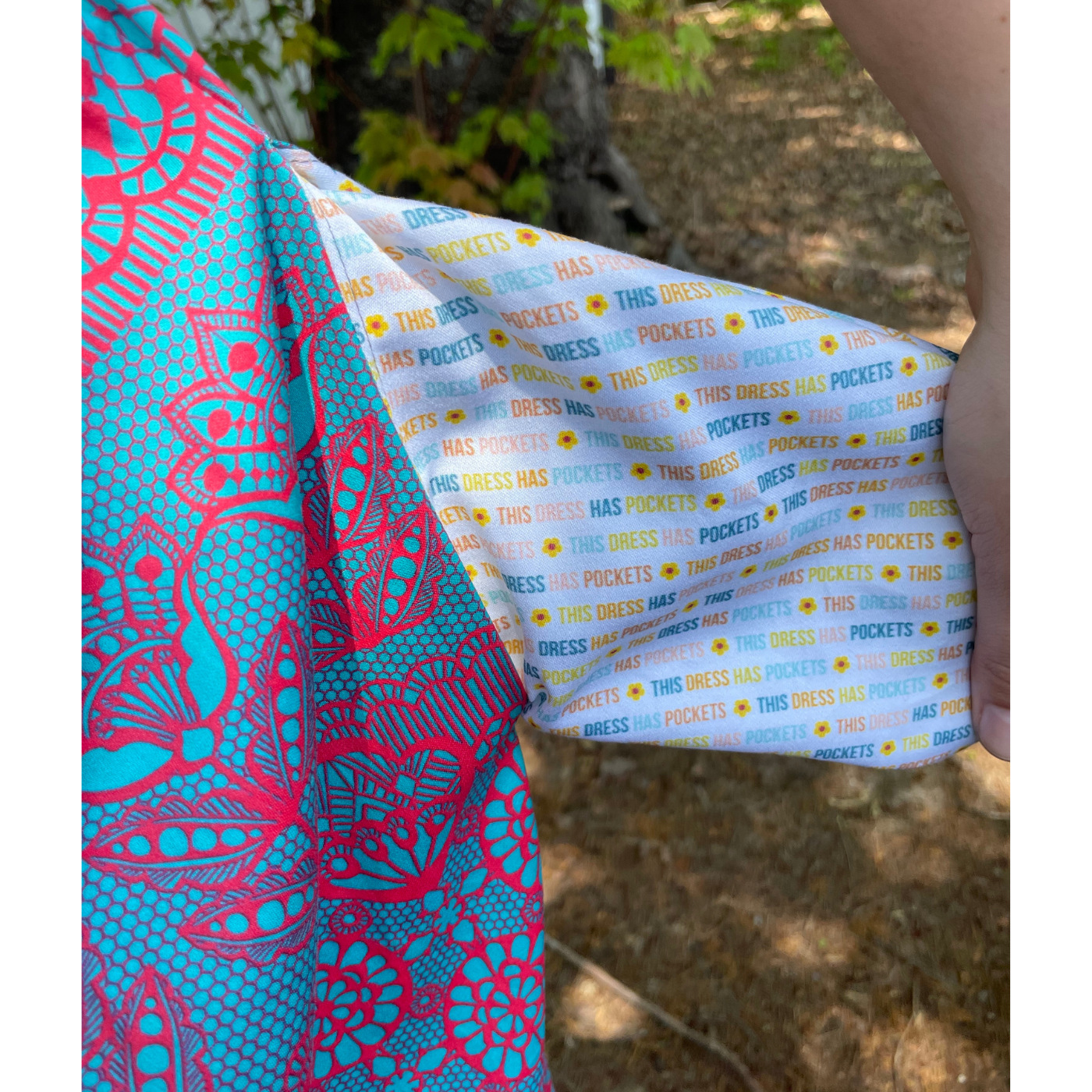 A close up of where a long turquoise dress with a hot pink lace design meets the pocket fabric. The pocket fabric design has a white background and small repeating text that says “This dress has pockets!” in turquoise, orange, yellow and blue with small yellow sunflowers between each repeating sentence.