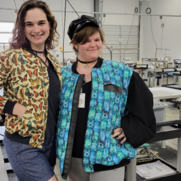 Sydney and Savvy stand in the Spoonflower factory in Durham wearing jackets that they made for Me Made May. Savvy wears a jacket with a zipper front. The jacket has small repeating columns of various zodiac signs in turquoise and teal blocks. Sydney is wearing jean shorts, a black shirt and jacket with black cuffs, a black zipper front and an orange, red and green butterfly design with a cream background.