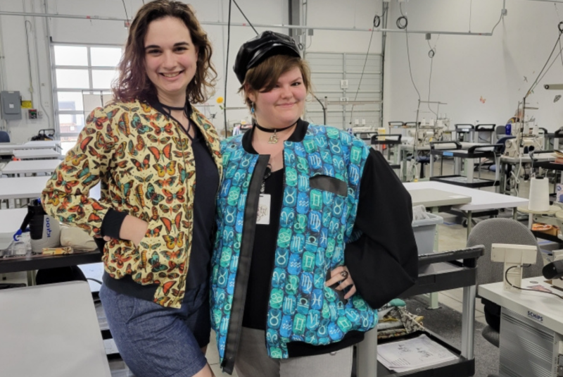 Sydney and Savvy stand in the Spoonflower factory in Durham wearing jackets that they made for Me Made May. Savvy wears a jacket with a zipper front. The jacket has small repeating columns of various zodiac signs in turquoise and teal blocks. Sydney is wearing jean shorts, a black shirt and jacket with black cuffs, a black zipper front and an orange, red and green butterfly design with a cream background.