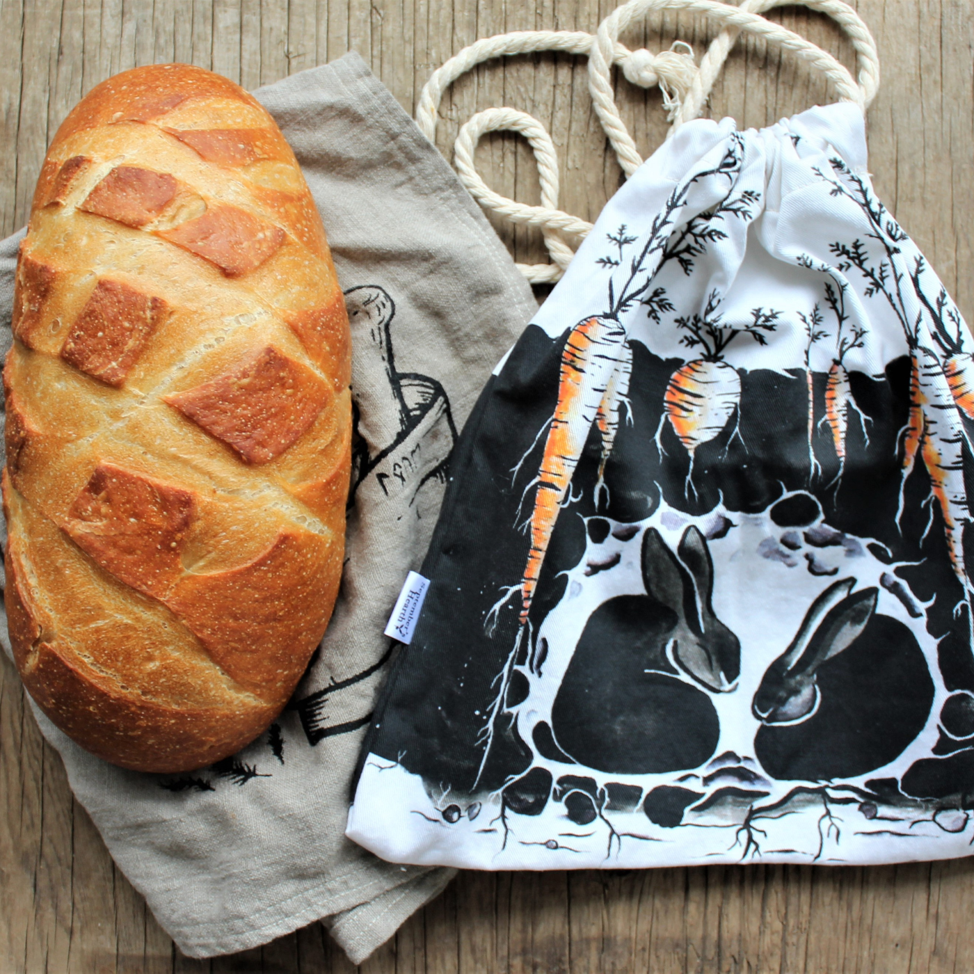 A white bread bag lays on a wooden table. The bag has a white rope closure and a design that peeks into a rabbit den showing two black rabbits curled up and sleeping with orange carrots growing in the earth above and on either side of them. A large loaf of bread is to the right of the bag. The bread sits on light brown fabric with parts of a black-lined design, that appears to be the edge of a hand-drawn mortar and pestle, showing on the right-hand side of the loaf.