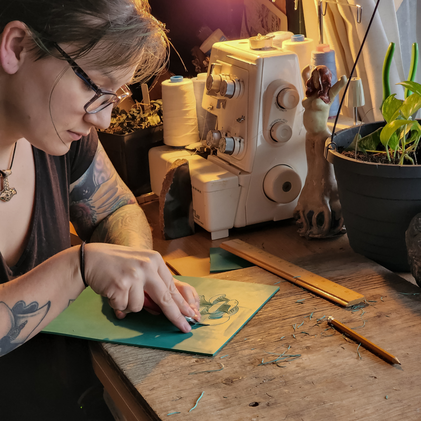 Sheena is leaning over a wooden table handcarving a stamp. A white serger, a dark green planter with leaves coming out of it, a wooden ruler and a pencil are also on the table.