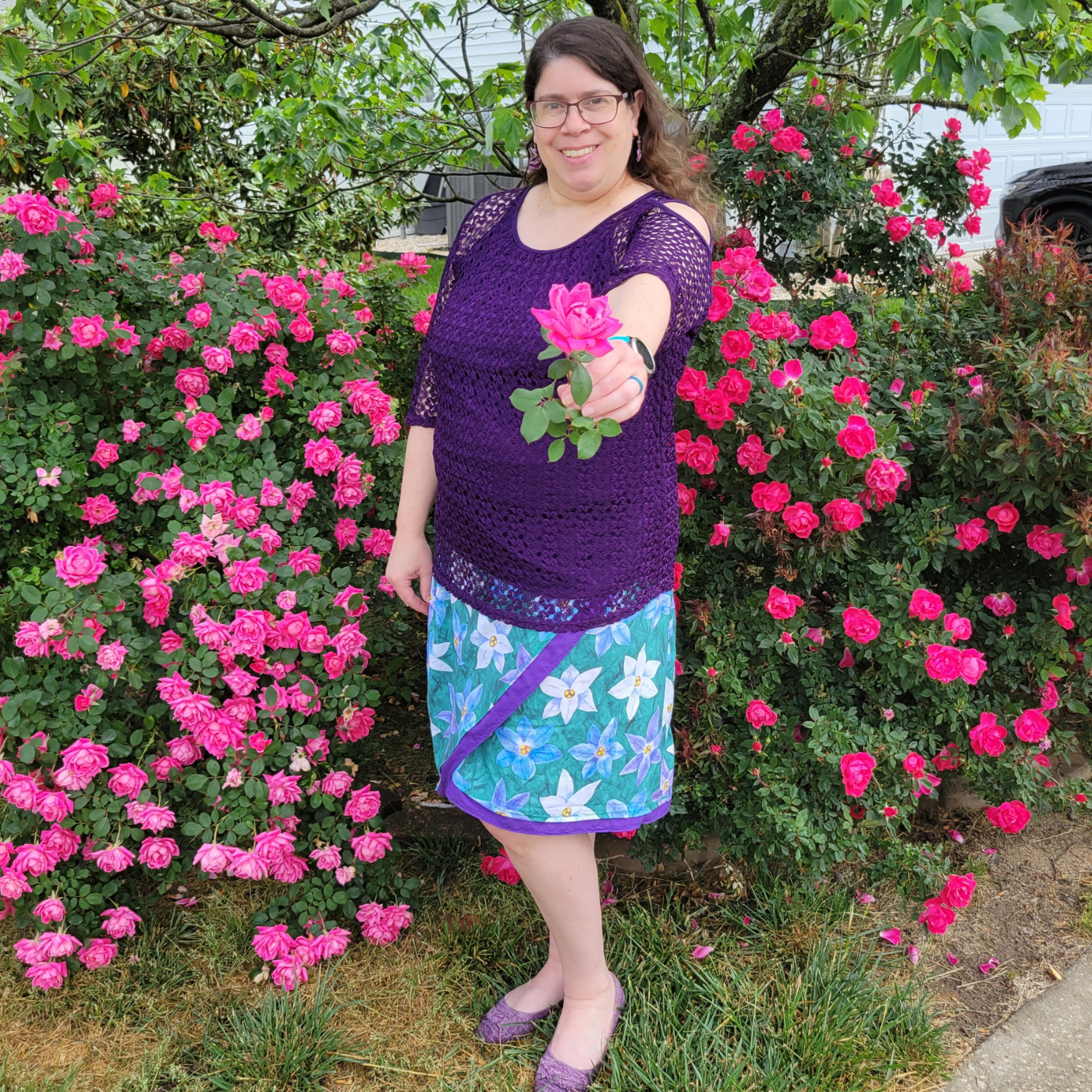 Sharon smiles and holds out a hot pink rose toward the camera. She is wearing a purple crochet top and tank top as well as a wrap skirt featuring a design with large purple, white and blue starflowers. The skirt has a strip of purple fabric along the hem and on the wrapped section part of the fabric, which goes diagonally down the front of the skirt.