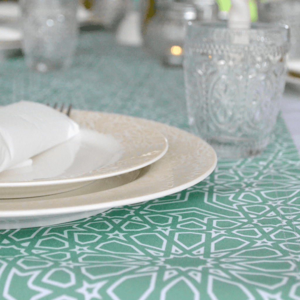 A large cream plate with a smaller plate on top of it, each with a tan floral design around their edges, sit on a table runner with a teal background and white geometric design laying on a white tablecloth. A knife and fork are rolled up in a white napkin on top of two plates. A short clear glass is to the top right of the plate, and other clear glasses are seen further down the table runner. 