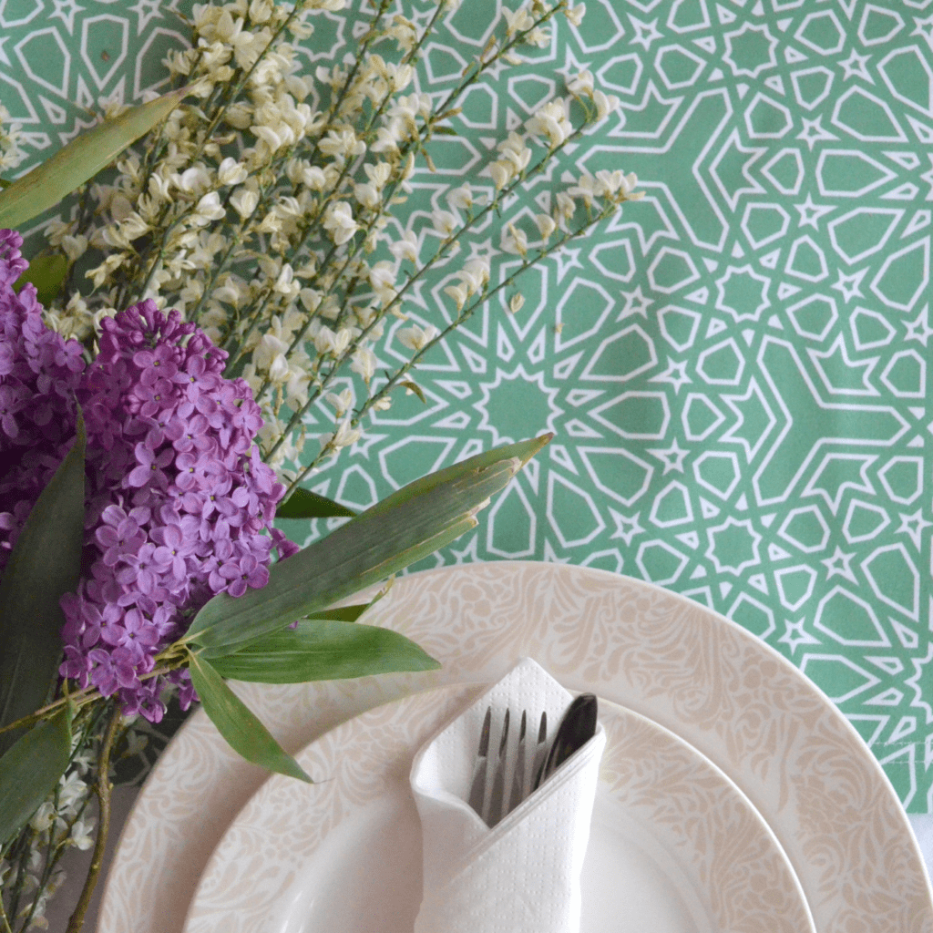 A large cream plate with a smaller plate laying on top sits on a table with a white tablecloth and a table runner with a teal background and white geometric design. A knife and fork are rolled up in a white napkin on top of the plates, which have a tan floral design around their edges. A bouquet with purple and white flowers and large green leaves lays on the table to the left of the plates. 