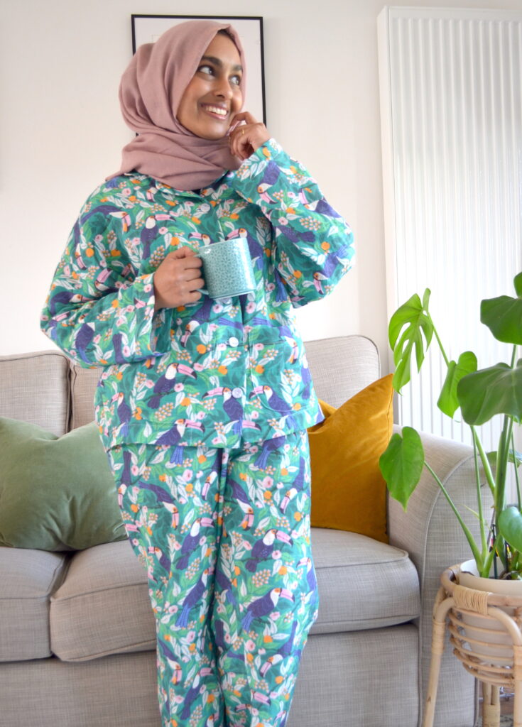 Rumana stands in a living room with a gray couch behind her; a green pillow is visible to her left and a dark yellow pillow is visible to her right. She is looking up and to the right and holding a blue mug. She is wearing a dusty pink hijab and pajamas with a tropical print featuring toucans, pink and orange flowers and a bright blue background. To the right of the photo is a monstera plant in a raised light bright wooden planter.