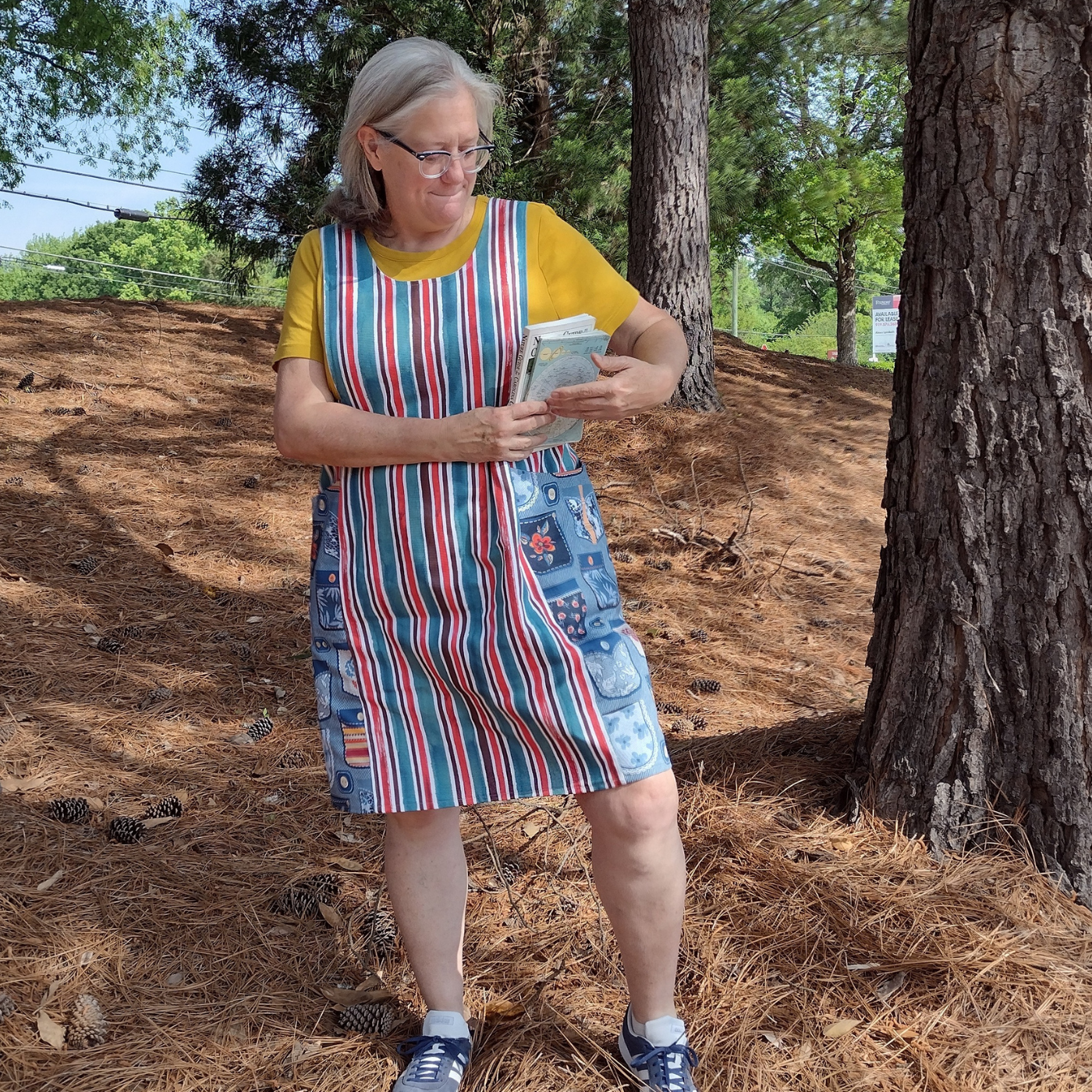 Sue stands in pine needles among a bunch of tall trees. She is wearing a red, blue and white vertical striped pinafore with large side pockets that have a design with a blue background and drawings of pockets on them. She is turning to the left and putting books in the leftside pocket.
