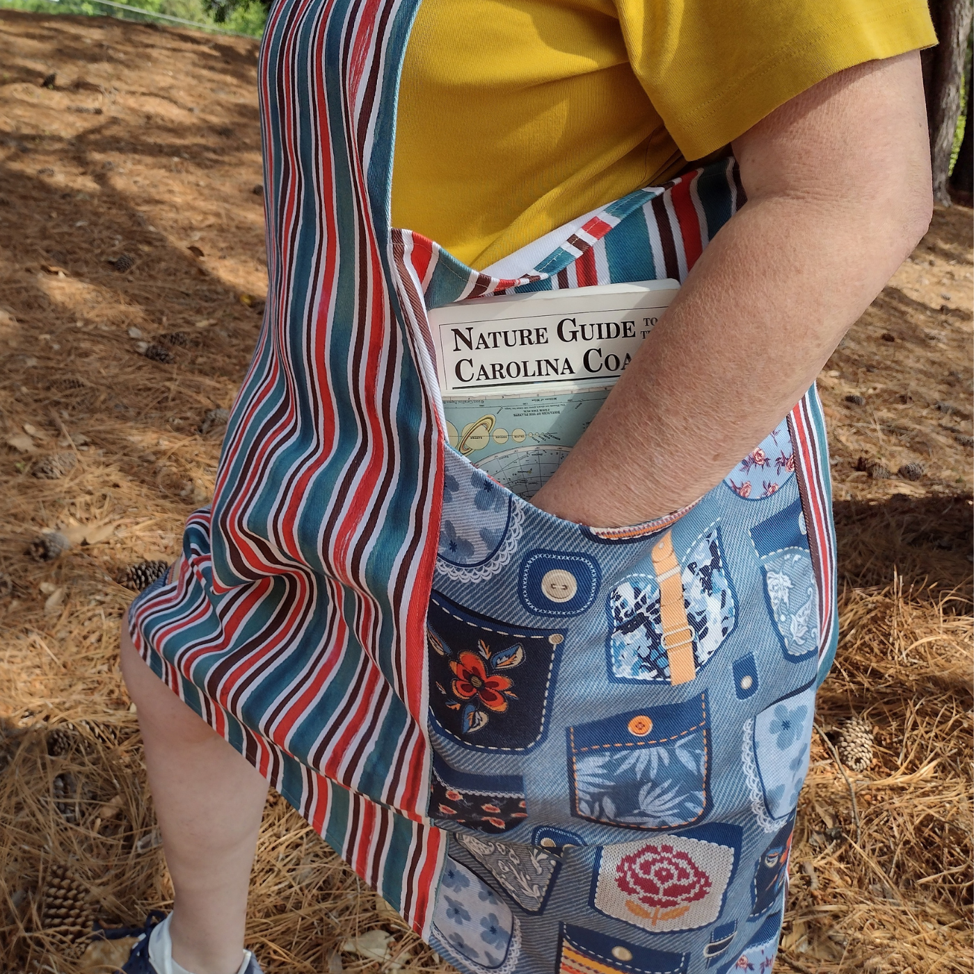 A close up of Sue’s red, blue and white vertical striped pinafore with large side pockets that have a design with a blue background and drawings of pockets on them. Her left arm is in the left pocket which has a book peeking out of that says “Nature Guide to the Carolina Coasts.”