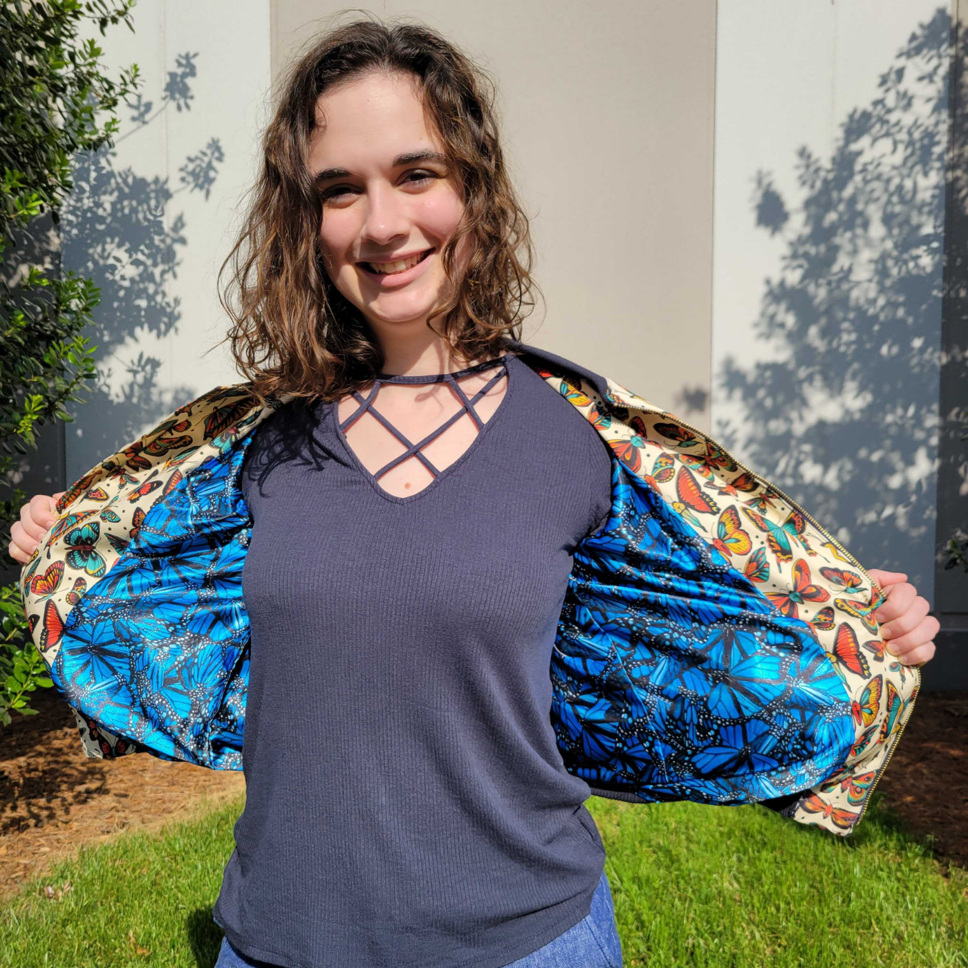 Sydney is wearing jean shorts, a black shirt and jacket with black cuffs and an orange, red and green butterfly design with a cream background. The middle sections of the butterflies are the lower half of human bodies as seen from behind. She is holding the jacket open to show a lining featuring a design filled with blue butterflies.