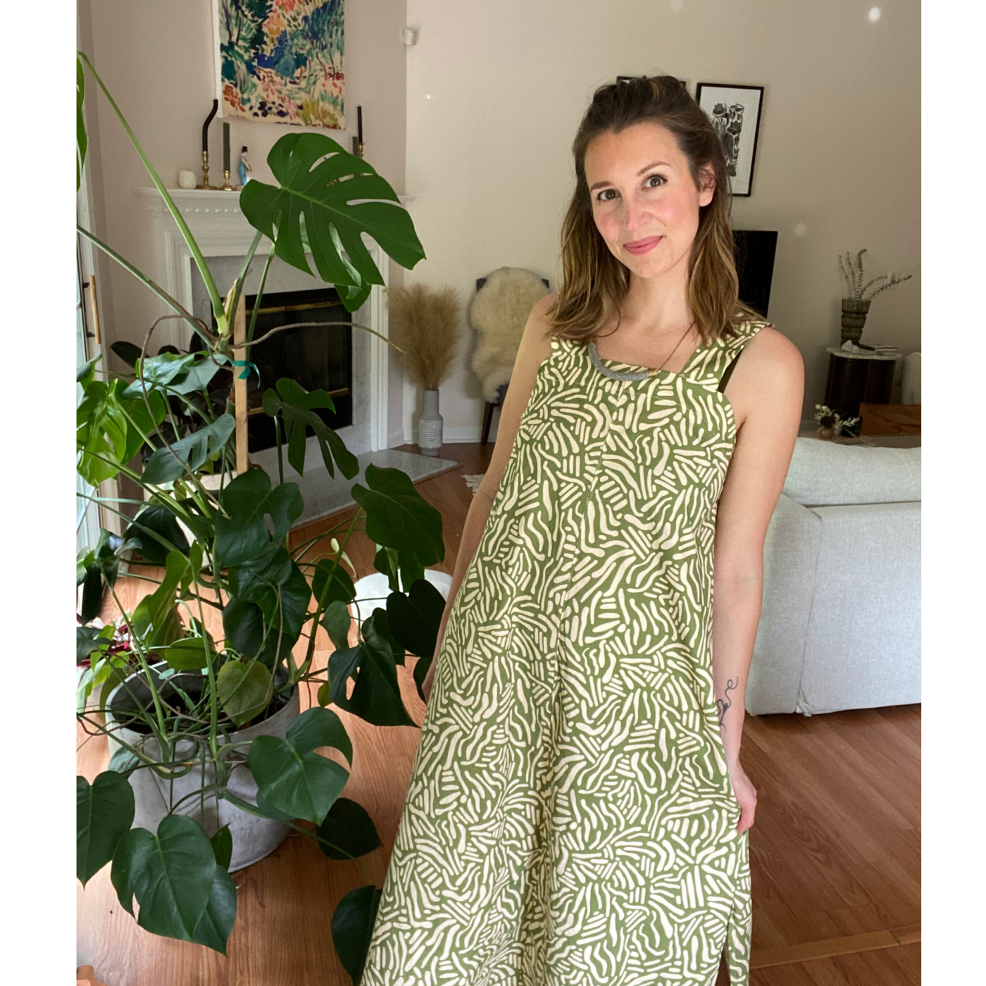 Theresa stands in a living room near a large monstera plant and smiles at the camera. She is wearing a long maxi dress with an olive green background and short wavy white lines running through the design in all directions.