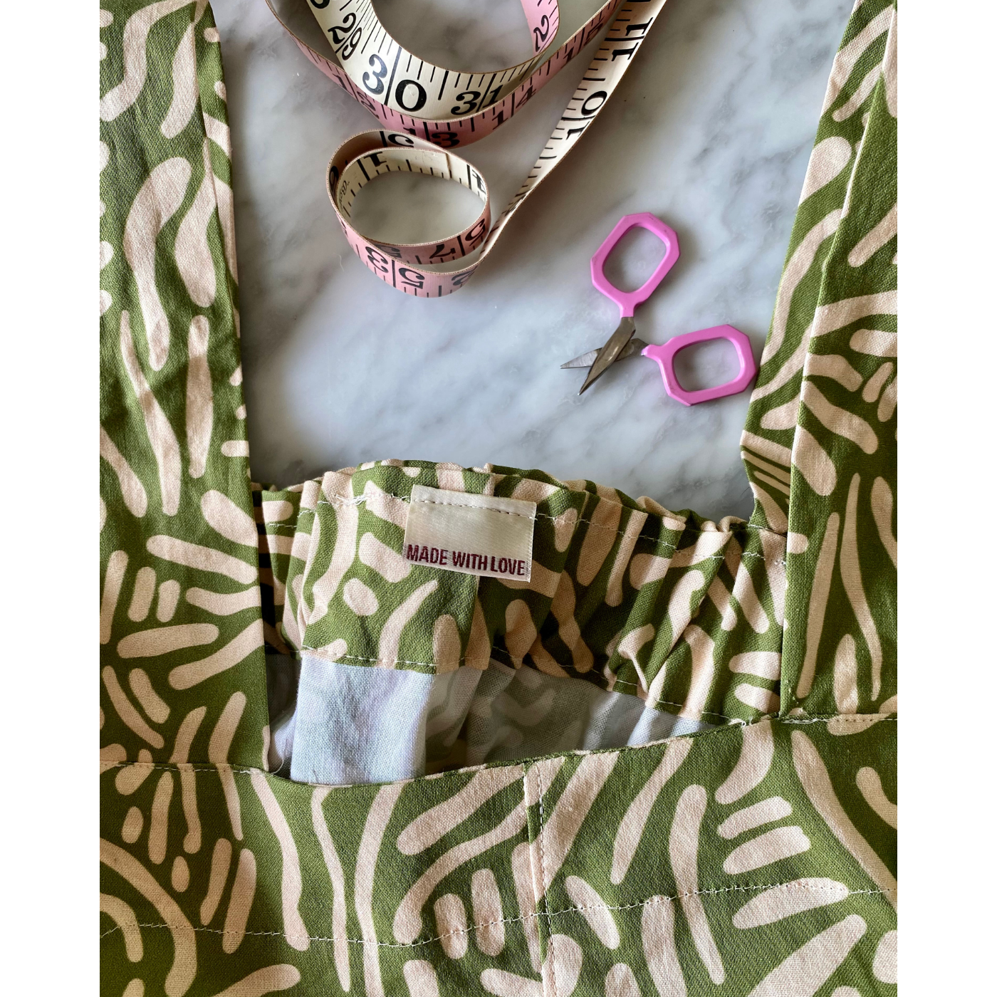 A close up of the inside neckline and top of a maxi dress laying on a white-and-gray surface with an olive green background and short wavy white lines running through the design in all directions. The neckline has a small tag that says “Made with Love.” A pink-and-white measuring tape and small pink scissors are on the table slightly above the dress.