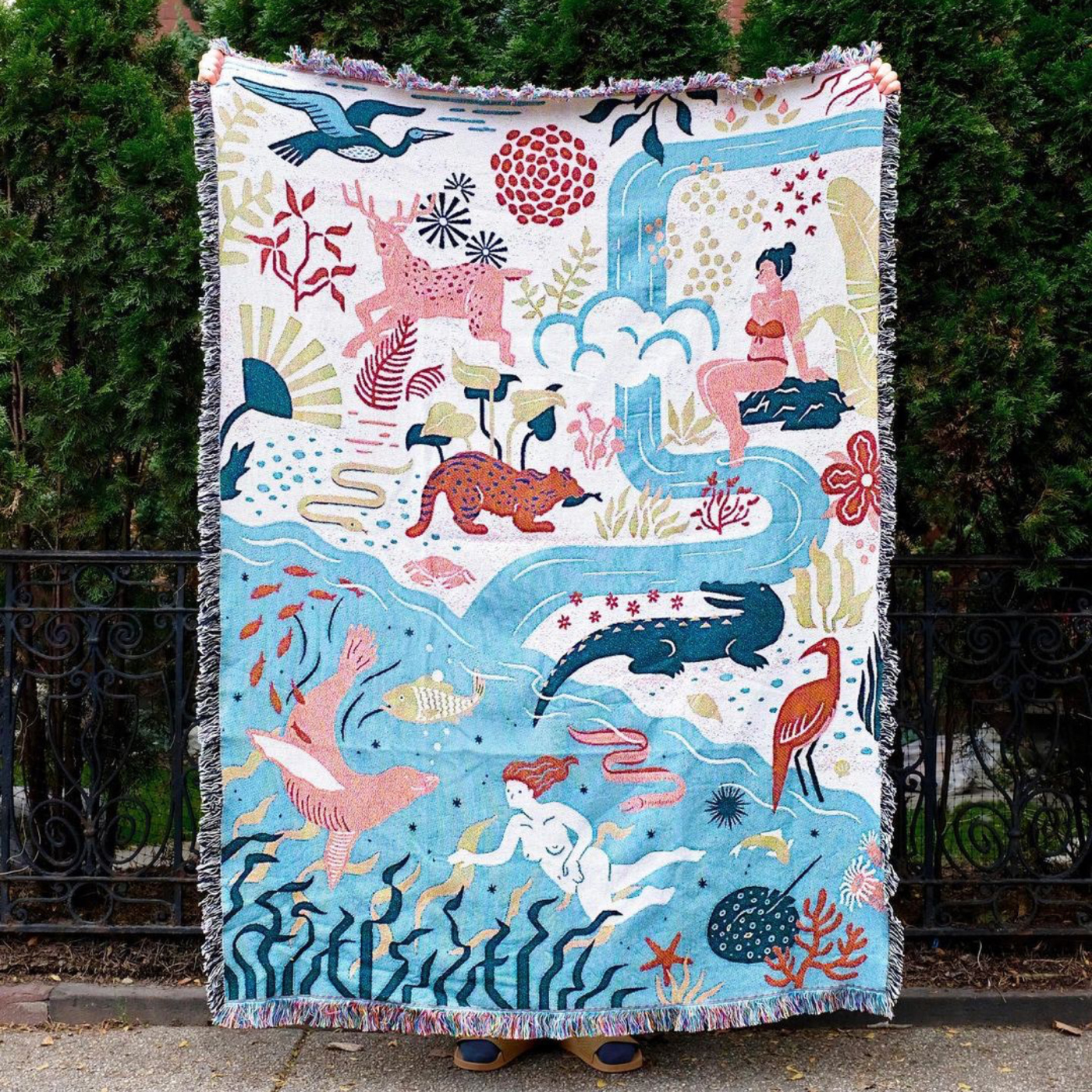 A throw blanket is displayed in front of some tall green shrubs. The top right half has a white background and the bottom left half has a blue background. The top right half features a person sitting on a rock in an orange bikini, a pink deer, a blue heron-like bird flying in the air, green branches, an alligator and more. A small stream starts in the top right-hand corner and pools down into the bottom left-hand side, where a mermaid swims with kelp, a seal, fish and small ocean wildlife.