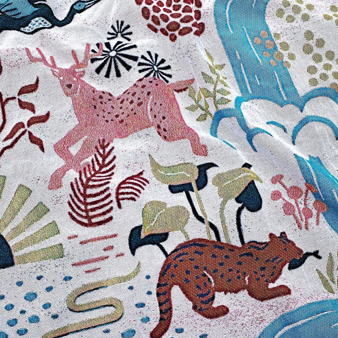 A close up of a throw blanket showing a section with a cream background, a pink deer, a stream of blue water coming from the top right side, a small brown animal with a black fish in its mouth, green leaves and more.