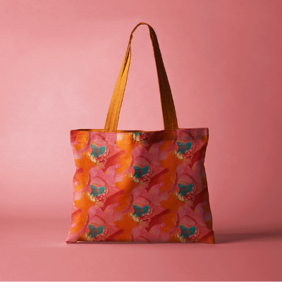 A tote bag with large abstract green, red and pink portions repeating on a red and orange background and orange straps. The sweatshirt is in front of a dusty pink background.