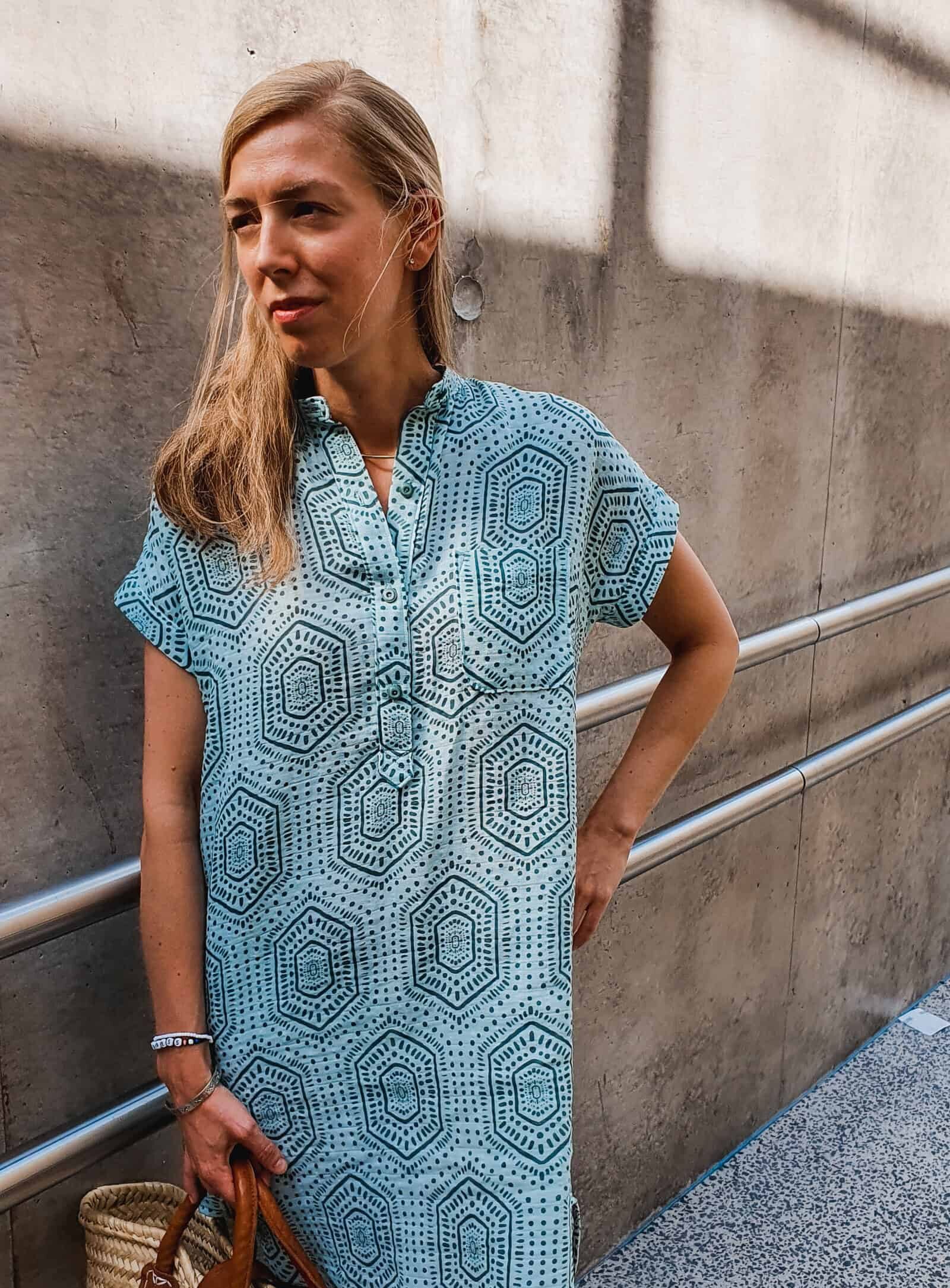 Clio Deprest in a blue gender-neutral long blouse in a beautiful boho style.  She is leaning in front of a wall.