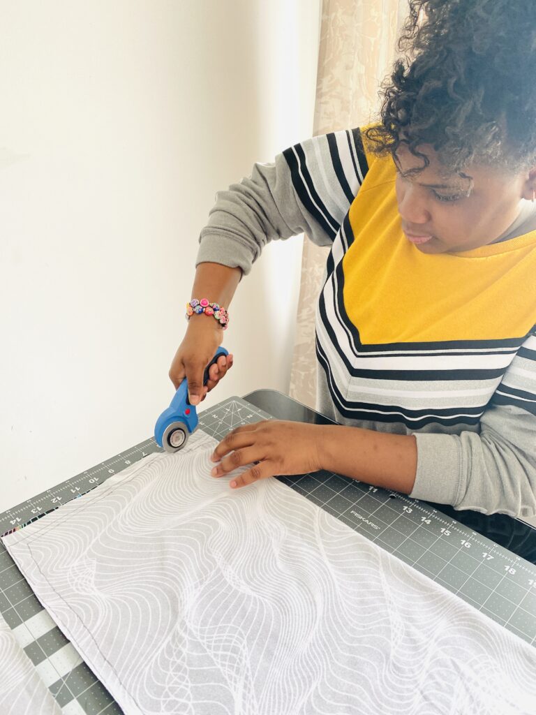 Ava uses scissors to cut out the body of a t-shirt, the wrong side of the fabric is showing, and the fabric is laying on a gray table. 