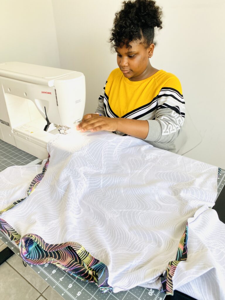 Ava sews the side seam of a t-shirt with the wrong side of the fabric facing up.  The right side of the shirt fabric is showing slightly at the seams and has a black background and blue, pink, yellow and purple holographic wavy lines all over it.