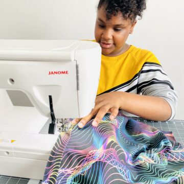Ava sews the neckband of a shirt to the body of the shirt with the wrong side of the fabric up. The right side of the shirt fabric is shown where the fabric has curled up, it has a black background and blue, pink, yellow and purple holographic wavy lines all over it.