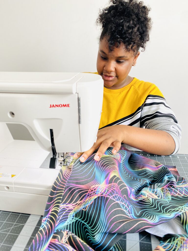 Ava sews the neckband of a shirt to the body of the shirt with the wrong side of the fabric up.  The right side of the shirt fabric is shown where the fabric has curled up, it has a black background and blue, pink, yellow and purple holographic wavy lines all over it.