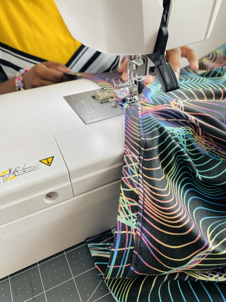 Ava sews the neckband of a shirt to the body of the shirt.  The shirt fabric has a black background and blue, pink, yellow and purple holographic wavy lines all over it.