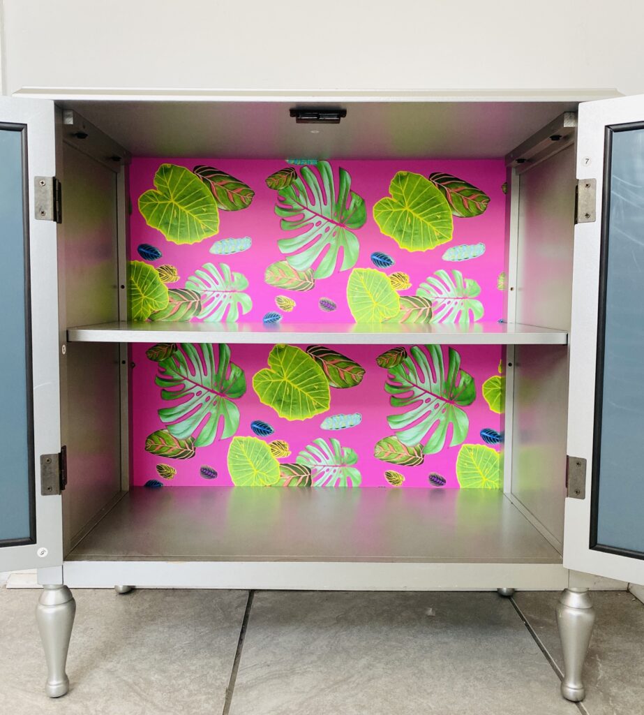 A small gray cabinet has its doors open showing that the inside back has been covered in peel and stick wallpaper featuring a design with plant leaves in different sizes and shapes on a hot pink background.  
