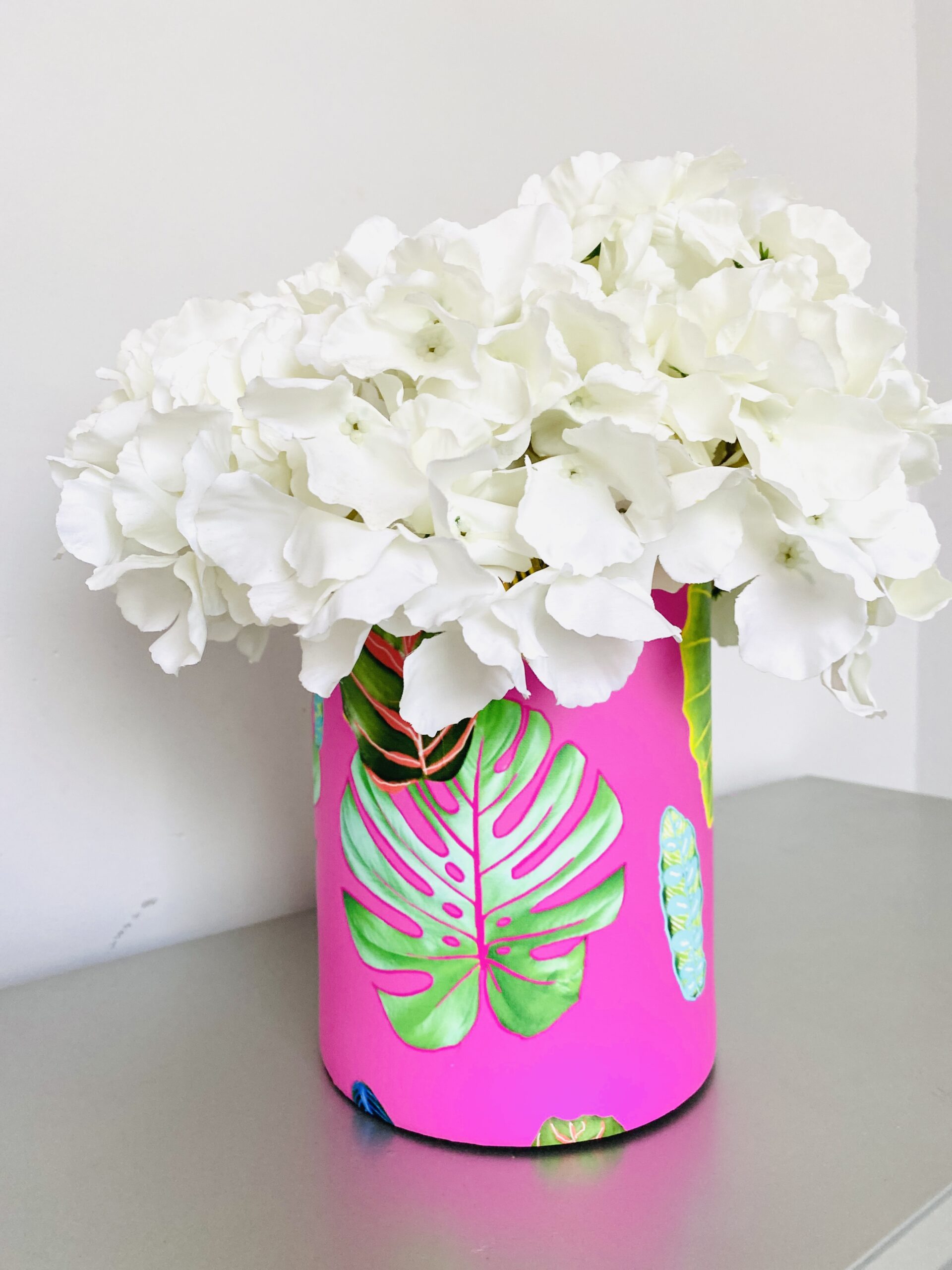 White flowers spill out of the top of a vase covered in peel and stick wallpaper. The design on the wallpaper features plant leaves in different sizes and shapes on a hot pink background. The vase sits on a silver surface and there is a wall white behind them.  