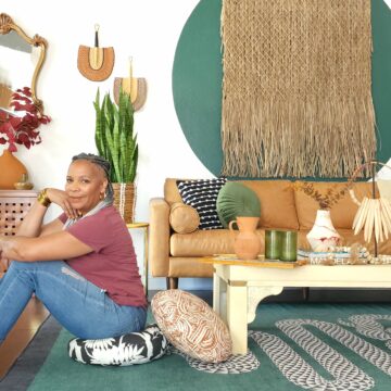 Tracey Hairston sits on a round floor cushion with a black background and white peonies floating throughout. A similar cushion with a rust background and white squiggly lines throughout sits behind Tracey. She sits in a living room with sage, light brown, white and cream accents. A sage carpet with a white-and-black geometric squiggle is on the floor, and a light brown leather couch is in front of a white wall with a circle painted sage in the center and a raffia wall hanging hanging in the middle of the center. The living room looks eclectic, luxurious and earth toned and filled with personal objects like a ceramic vase on a cream coffee table with dried flowers coming out from its top next to tall green glass glasses next to an earthen pitcher, to a snake plant in a woven container sitting in front of two small handmade fans hanging on the wall.