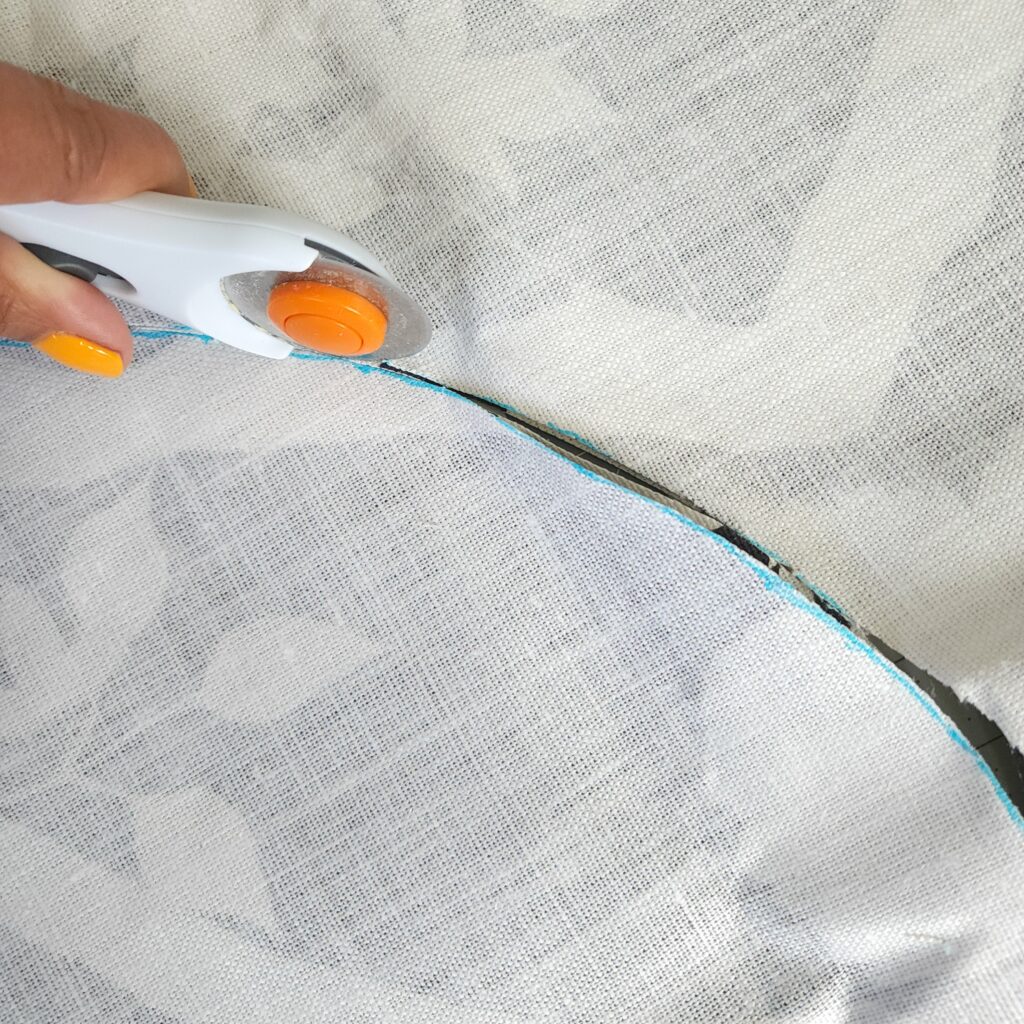 A hand holds a rotary cutter that is slicing through a blue line drawn on the wrong side of fabric as Tracey cuts out her cushion rounds.  