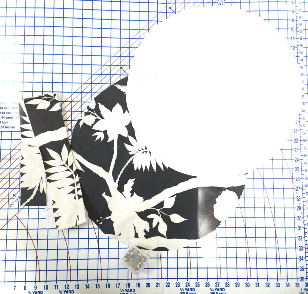 Three pieces of fabric featuring a design with a black background and white peonies lay on a white mat with a blue graph. The top of the cushion round is laying wrong side up. The bottom of the cushion round is lying right side up. The border strip of fabric lays to the left of the photo. A small clear box of pins lays at the photo’s bottom center. 