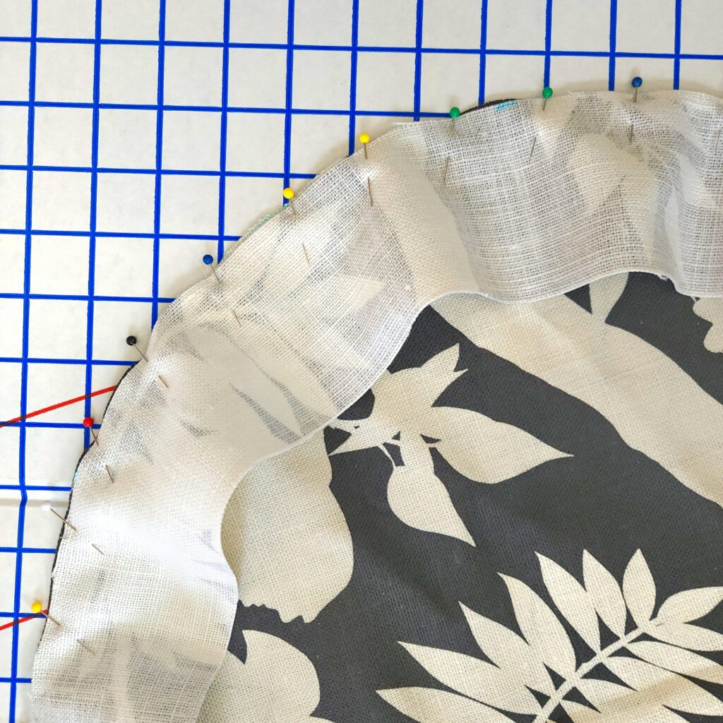 The wrong side of the border strip of fabric is shown pinned to a cushion round so that it can be attached. The fabric is laying on a white mat with a blue graph.
