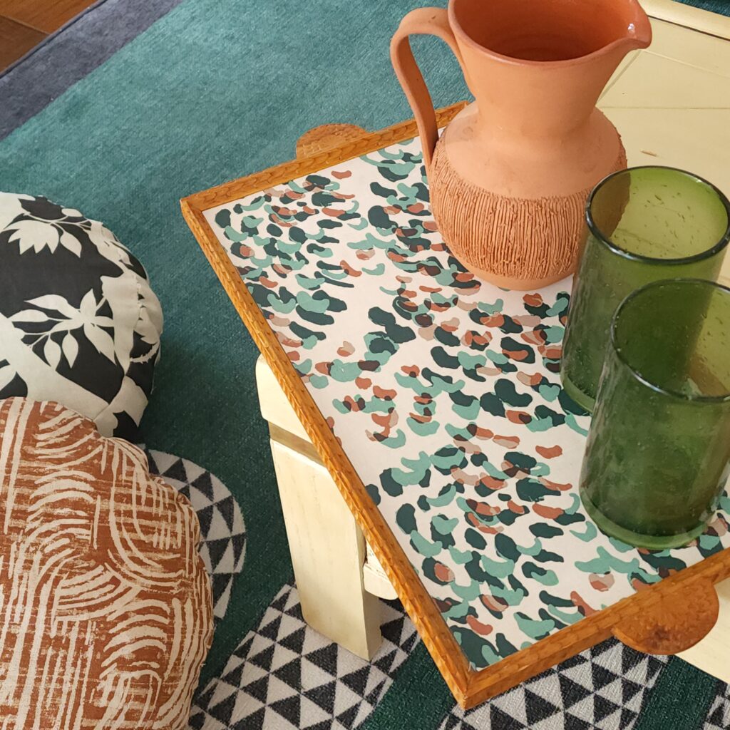 A wooden tray sits on a cream coffee table, the design on the tray has a white background and small brown, sage and dark green curved lines.  Two tall glasses made out of green glass sit next to an earthenware pitcher. 
