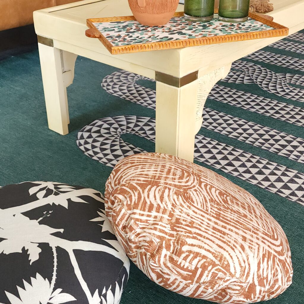 A wooden tray sit on a cream coffee table, the design on the tray has a white background and small brown, sage and dark green curved lines. Two tall glasses made out of green glass sit next to an earthenware pitcher. A sage carpet with a black-and-white geometric squiggle is on the floor. To the left of the coffee table are two stackable floor cushions, one with a black background and white peonies floating throughout and one with a rust background and white squiggly lines throughout.