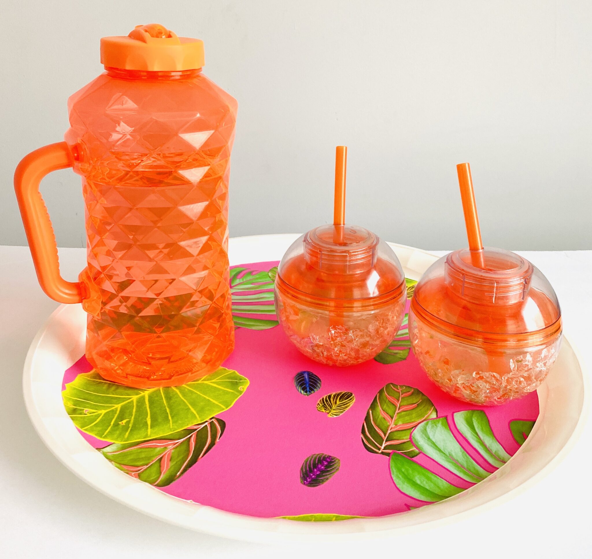 A plastic orange pitcher and two round orange cups with orange straws sit on a white serving tray on a white surface with white walls behind them. The design in the center of the serving tray features plant leaves in different sizes and shapes on a hot pink background. 