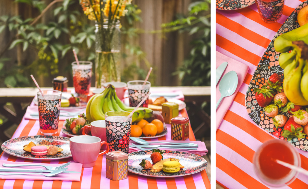 Two images side by side.  Close up of an outdoor table featuring both Spoonflower and Shutterfly designs.  The lilac-and-red thick stripe tablecloth and matching all-lilac napkins are from Spoonflower.  The plates, serving platter and plates from Shutterfly have a bright peach, orange, light yellow and red floral design with a black background.  Fresh fruit including clementines and bananas are placed on the serving platter in the middle of the table.  Pink mugs with red handles are also by each plate.  White forks, spoons and knives are placed on the napkins.  The table is surrounded by lush greenery.  A large clear glass vase is on the table with tall dried yellow and orange flowers.  On the right, a close up crop of the same table outdoor table with a focus on the tablecloth. 
