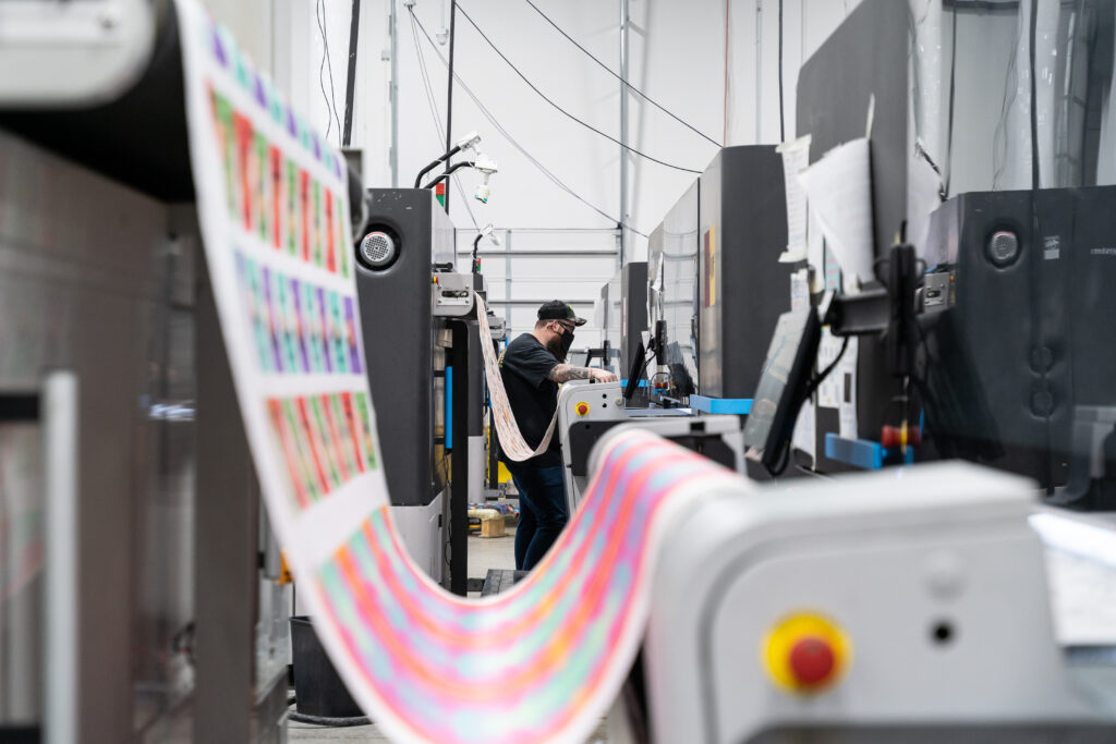 Several rows of tall digital printers are at work printing fabric in Spoonflower's Durham factory.  A person stands by the third machine in the row, showing a bit of scale as to the size of the printers, which are taller than the person. 