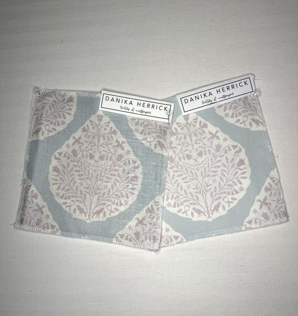 Small square swatches of a light purple floral paisley design on a light blue background lie next to one another on a white surface.  The photo is taken in incandescent lighting.  The swatch on the left has more of a blue textured background and the background on the swatch on the right is more flat and gray. 