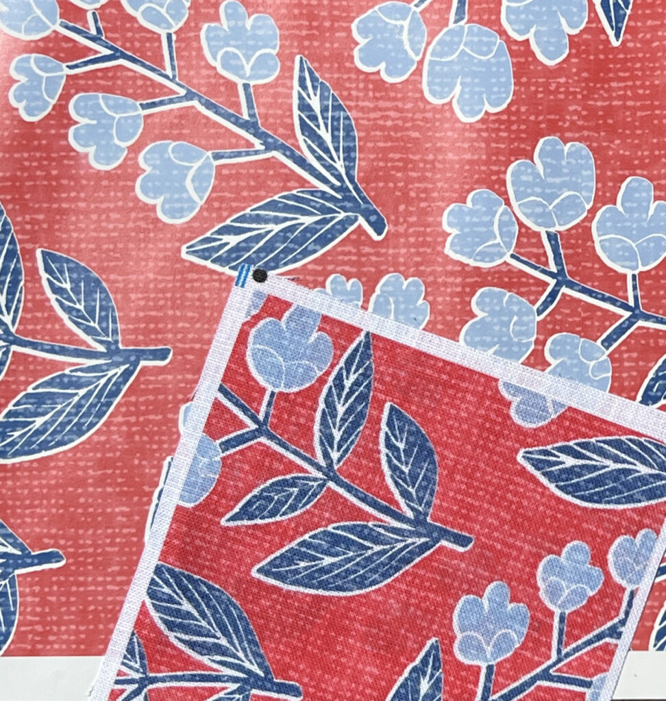 A square fabric swatch of a design with a dark red background and flowers with dark blue stems and light blue blooms lays on a swatch of wallpaper with the same design. 