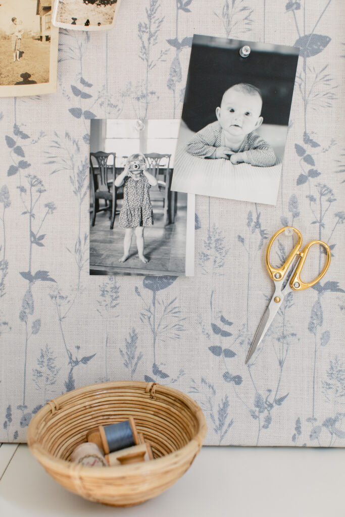   A close up of a fabric-covered bulletin board resting on the top of a low white bookshelf.  The fabric design has a white background and light blue flowers.  To the board's bottom right are two black-and-white photos of small children, one of an infant looking straight at the camera and one of a small child in a dress taking a photo and standing in front of a dining room table, and a pair of scissors with gold handles have been pinned to the board with clear push pins.  To the top left is a corner of a vintage black-and-white photo of a woman standing holding a baby next to a house.  On the bookcase in front of the bulletin board is a small wooden nest with several spools of thread inside. 
