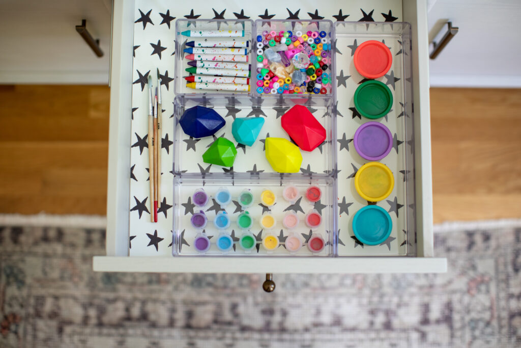 A small white drawer is shown open and from above.  A neutral geometric carpet and brown floor are below it.  The drawer liner is made of peel-and-stick wallpaper with a white background and rows of small black stars.  The drawer contents are children's art supplies, from left to right, three small wooden paintbrushes, a clear four-part open storage container with gem-shaped crayons, stick-shaped crayons, colorful beads and small pots of paints.  To the right is a stack of vertically arranged pots of modeling compound. 