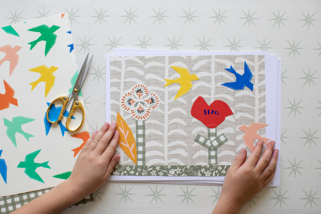 Two hands are on either side of a piece of paper as seen from above.  The paper is laying on a scrap of white wallpaper with gray stars and has a nature scene on it crafted out of wallpaper scraps: a red flower, a blue bird, a yellow bird, a tree made from small wallpaper scraps of flowers stuck together on a tan background with thick white-lined trees.  To the left of the paper are a pair of scissors on top of a scrap of wallpaper with a white background and yellow, pink, green, orange and blue birds. 