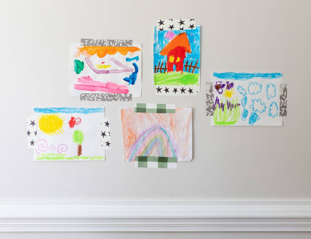 Five pieces of children's art are grouped together on a white wall, taped to it with small rectangular wallpaper scraps.  All artwork is on a white piece of paper, clockwise from top left: A piece of artwork with several squiggles in bright colors is taped to the wall with scraps with a gray background and white stems;  a red house with a brown picket fence and pink chimney is taped to the wall with scraps made of black stars on a white background;  geometric squiggles are taped to the wall with scraps featuring a gray background, white cheetahs and white stems;  a rainbow against a peach sky is taped to the wall with green-and-white gingham scraps;  and a piece with a red bird under a blue sky flying away from a large yellow sun and over a green tree is taped to the wall with scraps made of black stars on a white background.