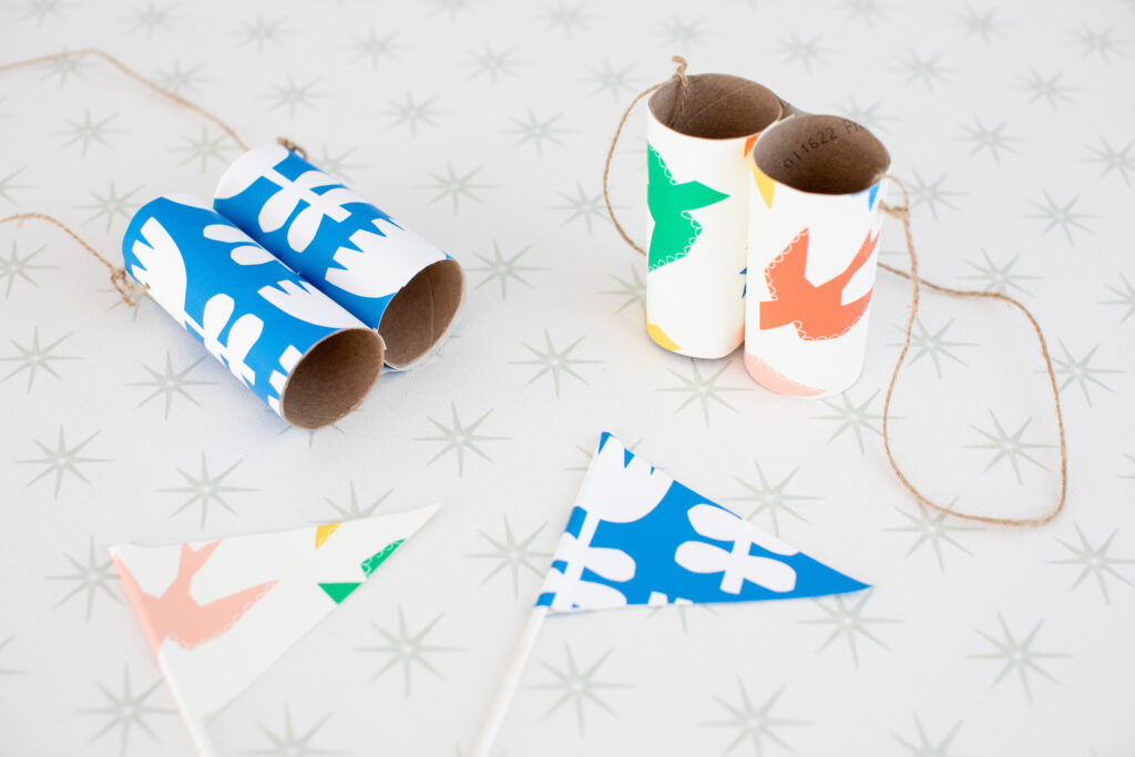 Wallpaper scraps have been used to create items for kids' adventure and lay on a scrap of white wallpaper with gray stars.  Wallpaper with yellow, green, red and blue birds on a white background covers a pair of toilet paper roll binoculars and small pennant flags.  A pair of toilet roll binoculars is also covered in blue wallpaper with large white flowers. 