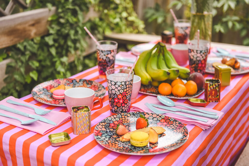   Photo of an outdoor table featuring both Spoonflower and Shutterfly designs.  The lilac-and-red thick stripe tablecloth and matching all-lilac napkins are from Spoonflower.  The plates, serving platter and plates from Shutterfly have a bright peach, orange, light yellow and red floral design with a black background.  Fresh fruit including clementines and bananas are placed on the serving platter in the middle of the table.  Pink mugs with red handles are also by each plate.  White forks, spoons and knives are placed on the napkins.  The table is surrounded by lush greenery. 