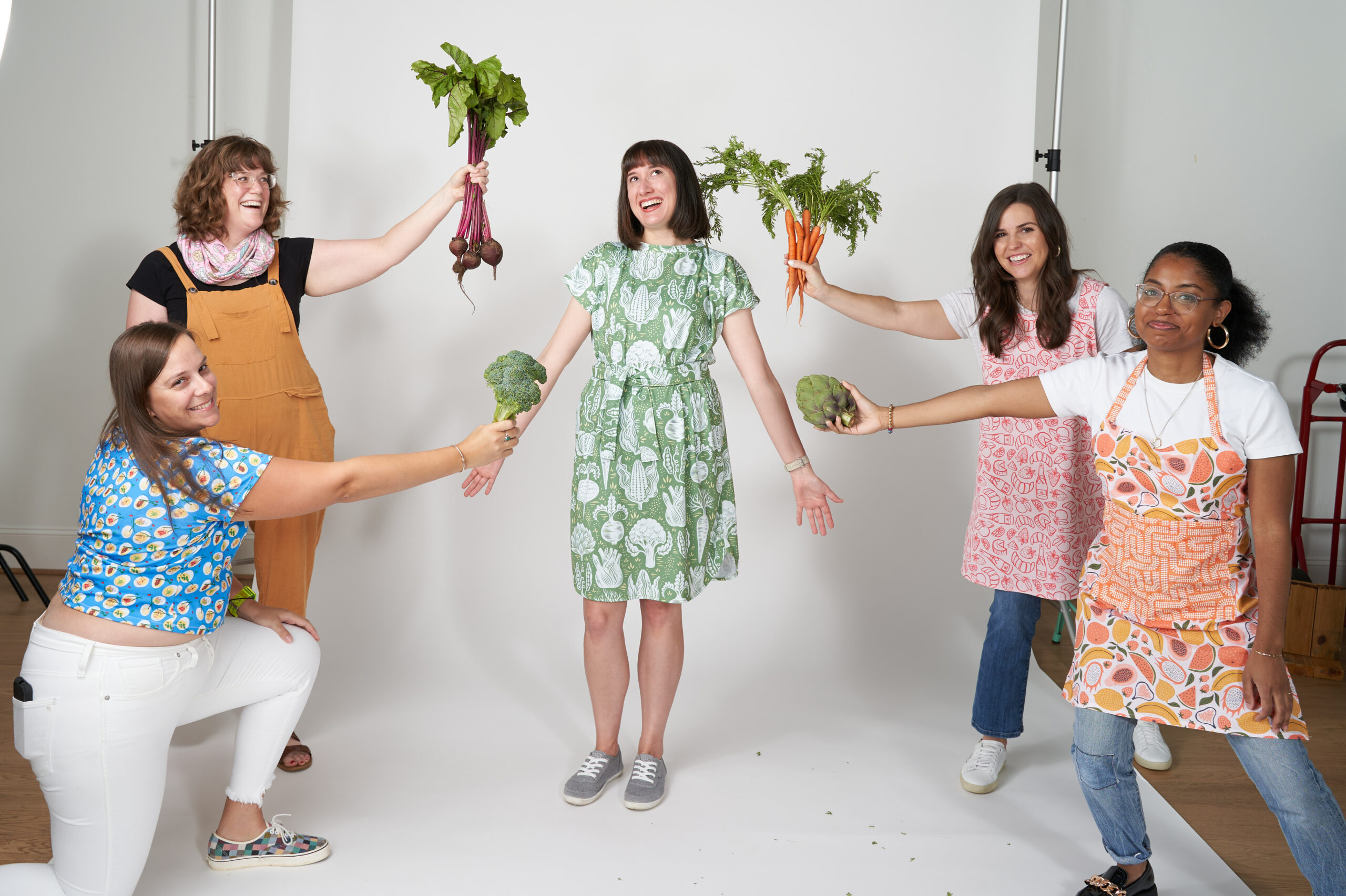 A group of women stand in front of a white back drop in a photo studio holding up various vegetables around a woman in the center wearing a vegetable dress