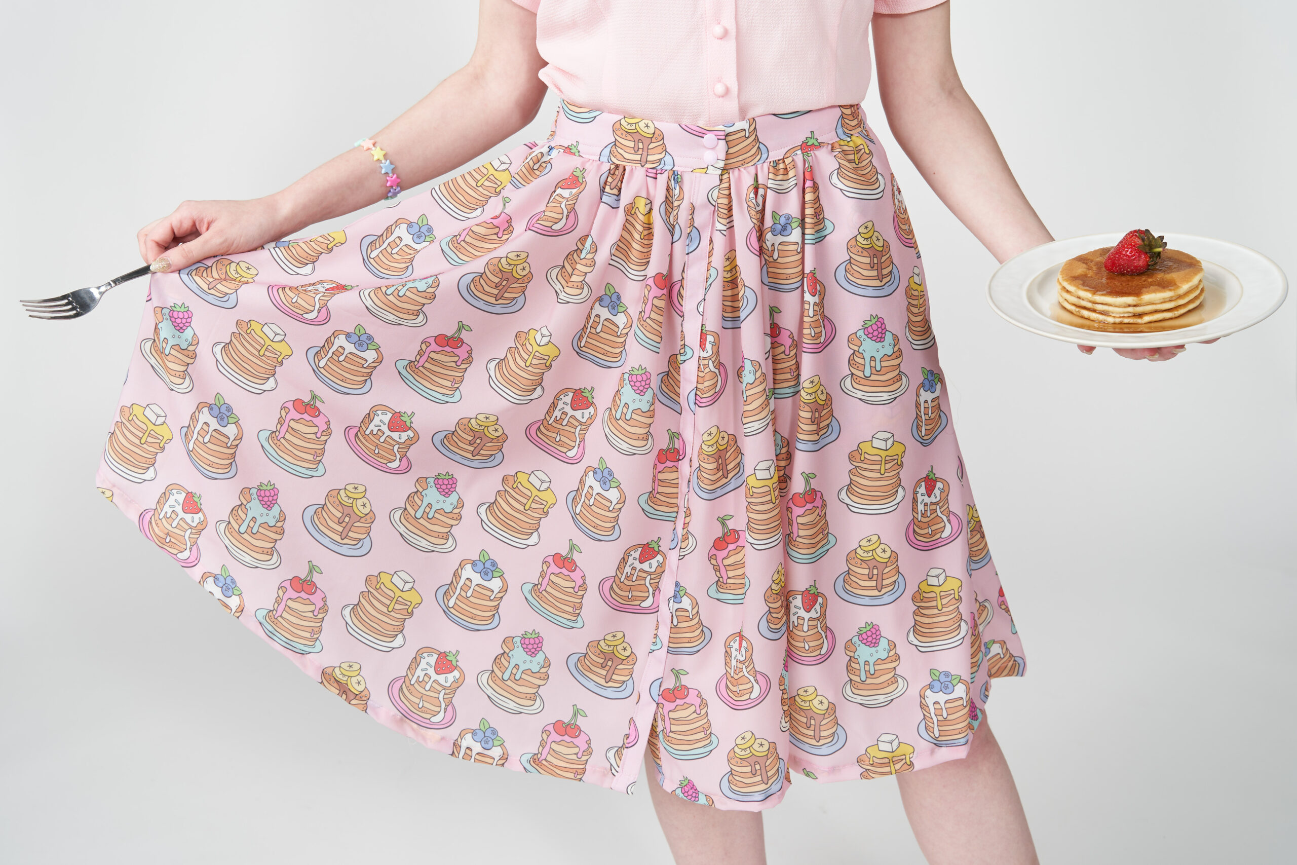 Anna is a white woman with long reddish blonde hair wearing a pink button up blouse and pink pancake button up skirt. She is holding a plate of pancakes with syrup and strawberry.