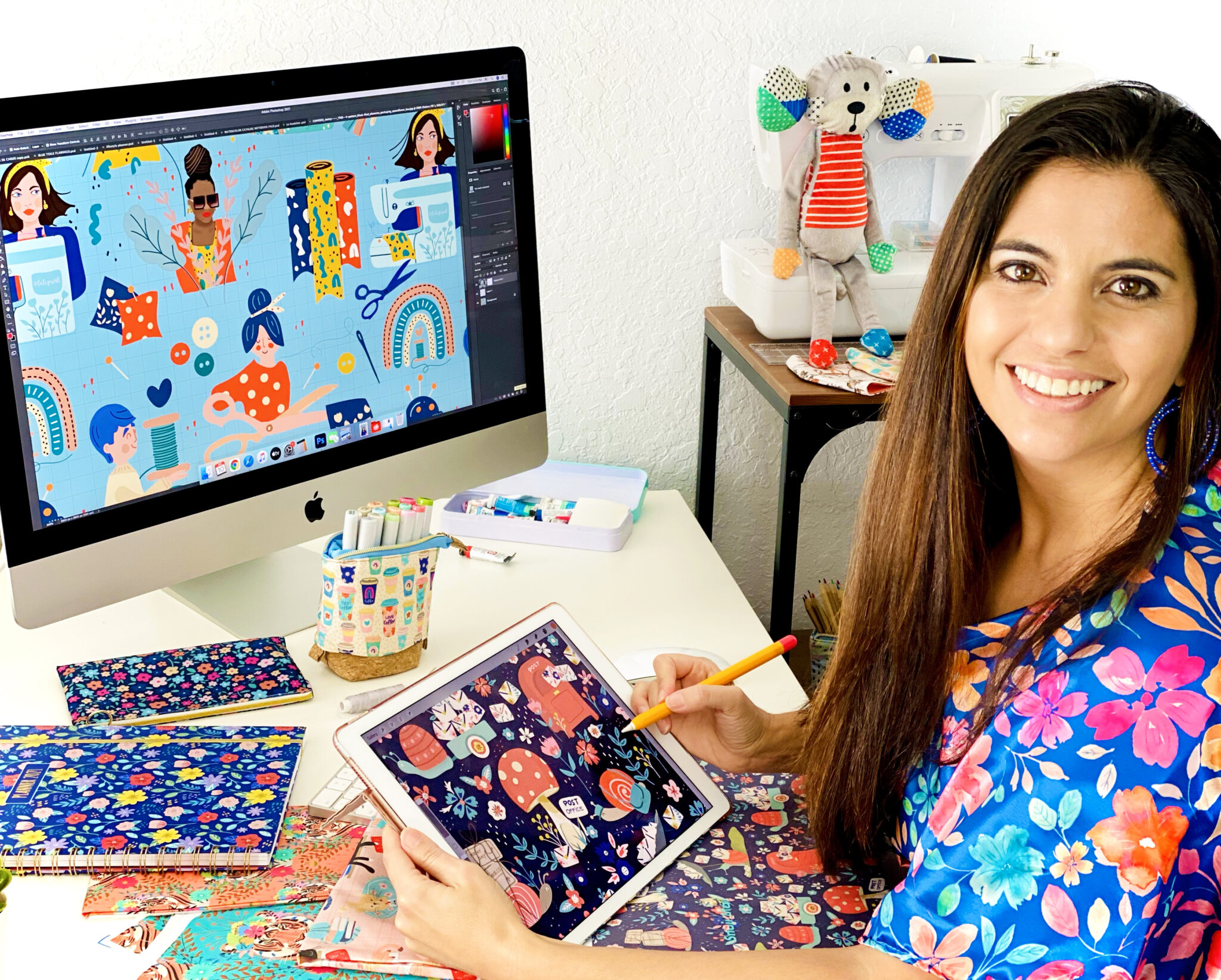 Luciana is sitting at a computer desk and turns toward the camera smiling. She is drawing on a tablet on the desk with a large monitor in front of her. The monitor, the tablet, notepads and stationery on the table are all bright, cheery florals, some with people making things or drawn with craft supplies and some with animals, like snails or tigers.