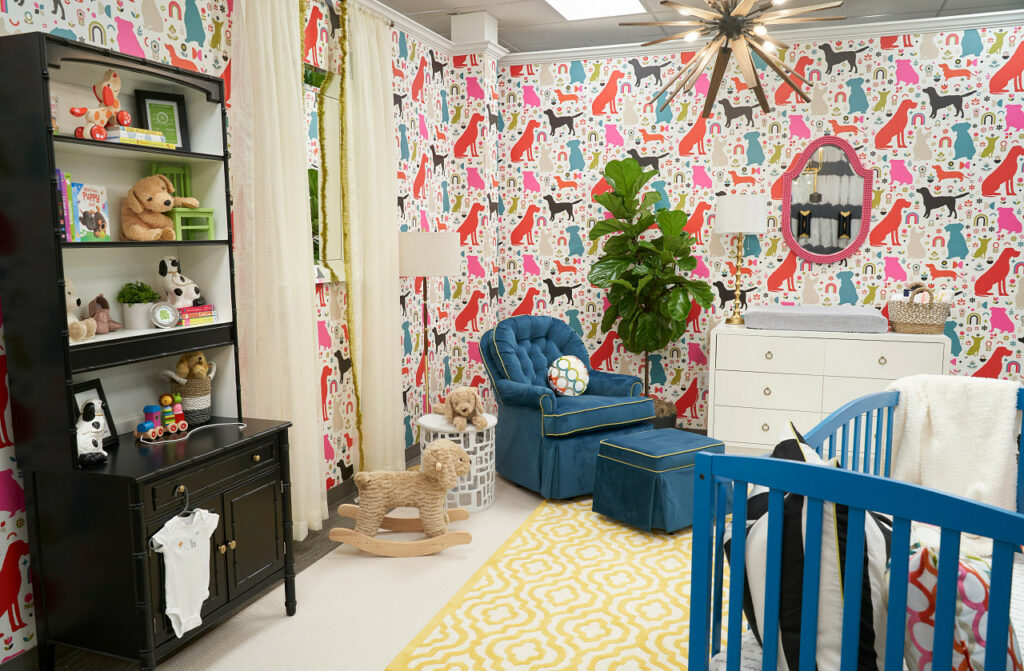 A nursery with red, pink, green and blue wallpaper patterned with mid-century dogs. A blue crib matches the blue velvet chair. There is a black bookshelf with stuffed dogs and books.